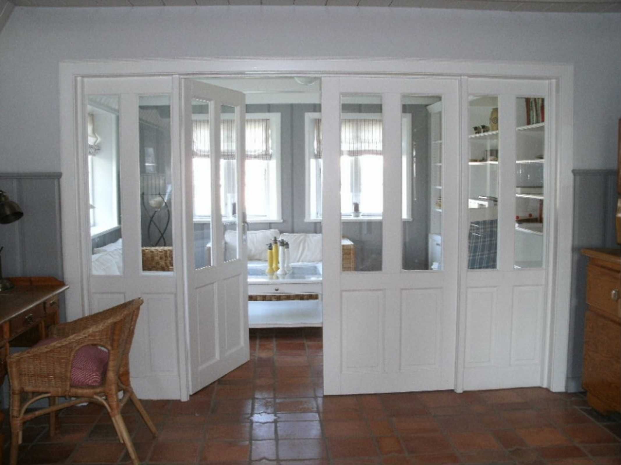 A room with a white door leading to a bedroom, a wicker chair, a tiled floor, and a bookshelf.