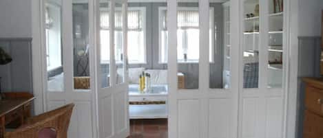 A room with a white door leading to a bedroom, a wicker chair, a tiled floor, and a bookshelf.