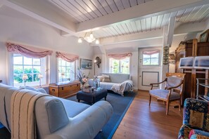 A cozy living room with a sofa, ottoman, and armchair, wooden flooring, and a large window with curtains. - Anno 1631, Anne Bonney No. 2, Wing (Süderende)