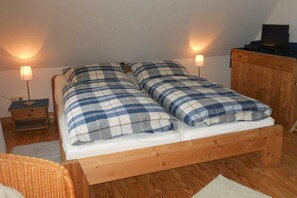 A small, cozy bedroom with a wooden bed, two bedside lamps, a wooden dresser, and a wicker chair.