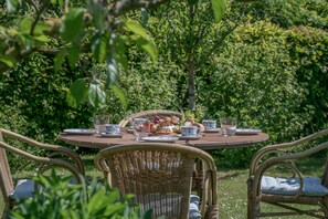 Une table ronde en bois, dressée pour un repas, entourée de chaises en osier et d’une végétation luxuriante.