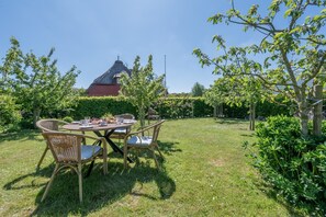 Ein Essbereich im Freien mit Korbstühlen und einem Tisch für zwei Personen, umgeben von üppiger Vegetation und einem roten Gebäude im Hintergrund.