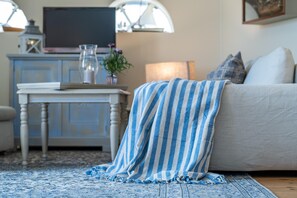 Ein gemütliches Wohnzimmer mit einer gestreiften Decke über der Couch, einem kleinen Tisch mit Buch und Vase sowie einem Fernseher auf einem Schrank.