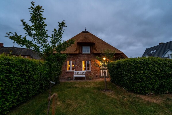 Une maison avec un toit de chaume, un banc blanc dans le jardin et une haie bien entretenue.