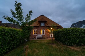 Une maison avec un toit de chaume, un banc blanc dans le jardin et une haie bien entretenue.