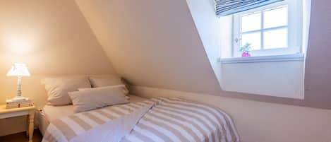 A bedroom with a sloped ceiling, a window with a view, a bedside table with a lamp, and a bed with striped bedding.