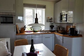 A compact kitchen with a dining area, featuring white cabinets, a built-in oven, a hanging light fixture, and a window providing natural light.