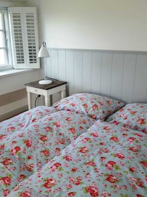 Une chambre à coucher avec un lit recouvert d’une literie à motifs floraux, une table de chevet avec une lampe et une fenêtre avec des volets.