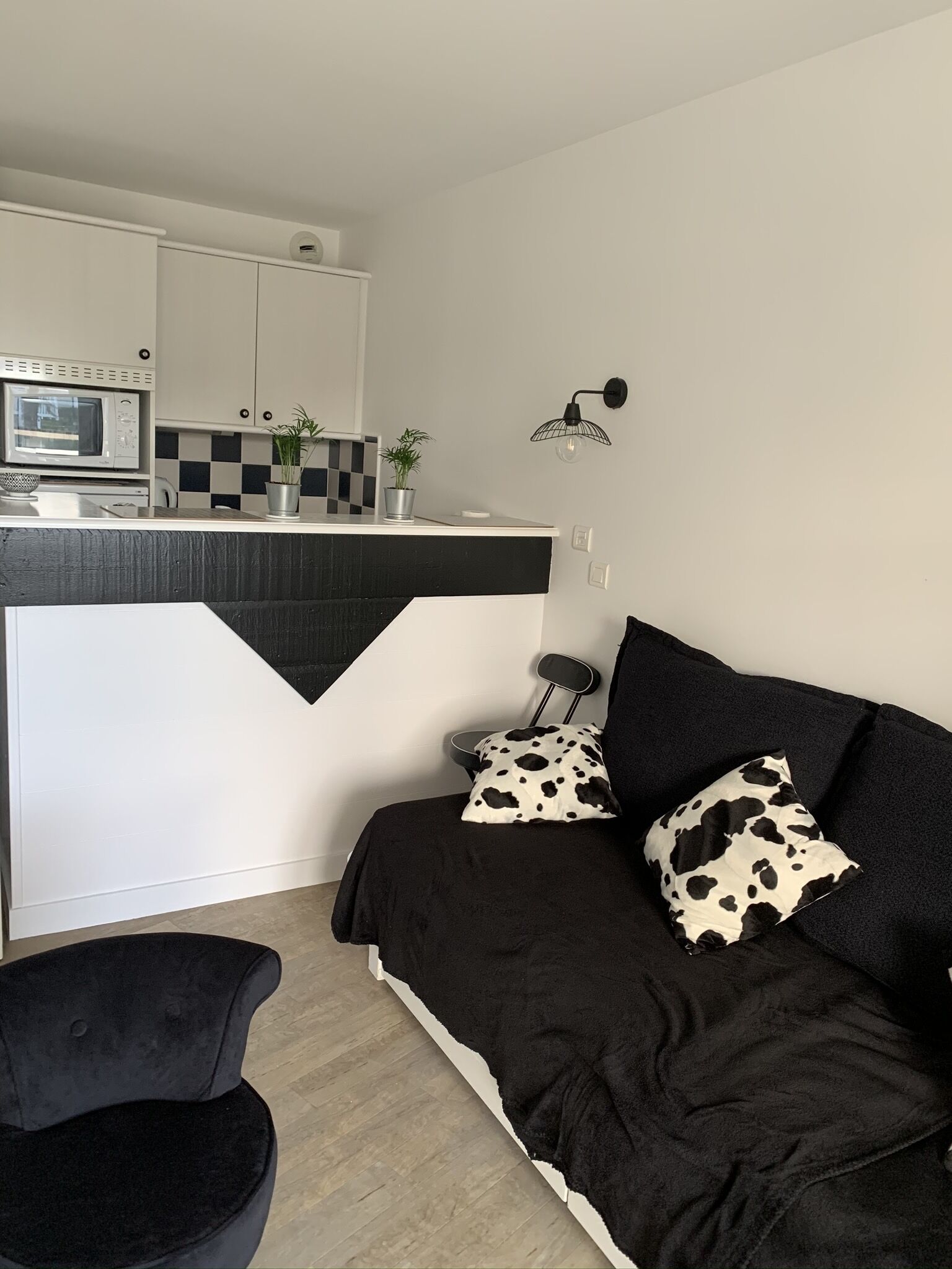 Un salon moderne avec un canapé noir et blanc, un fauteuil noir et un coin cuisine équipé d’un four à micro-ondes et d’un crédence à carreaux.