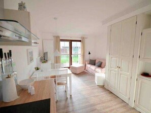 A spacious room with a large window, a dining area with a white table and chairs, a sofa, and a wardrobe. - Kliffkoje, Guatingwai 12 Foehr (Nieblum)