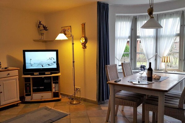 A living room with a television, a dining table with chairs, a floor lamp, and a window with curtains.