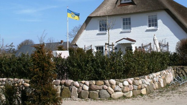 Een wit huis met rieten dak, een stenen muur ervoor, en een Oekraïense vlag.