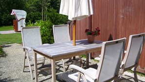 Een terras buiten met een tafel en stoelen, een witte parasol en een bloempot met rode bloemen.