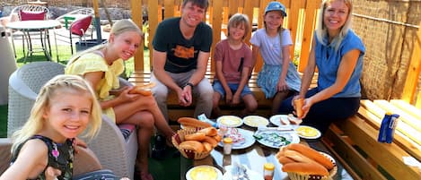 Un groupe de personnes est assis autour d’une table en bois, devant de la nourriture et des boissons, à l’extérieur, avec des bâtiments visibles en arrière-plan.