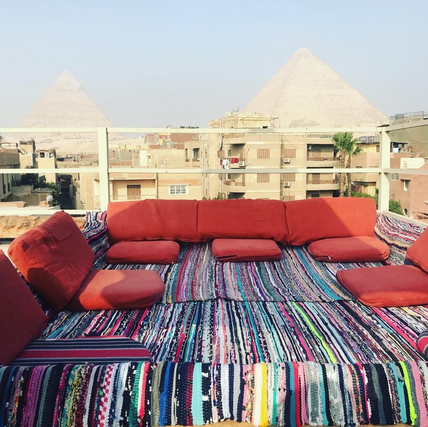 Un espacio de estar en la azotea con un tapete a rayas de colores y cojines rojos, con vistas a un paisaje urbano que incluye edificios y pirámides al fondo.