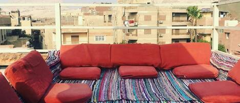 Un espacio de estar en la azotea con un tapete a rayas de colores y cojines rojos, con vistas a un paisaje urbano que incluye edificios y pirámides al fondo.