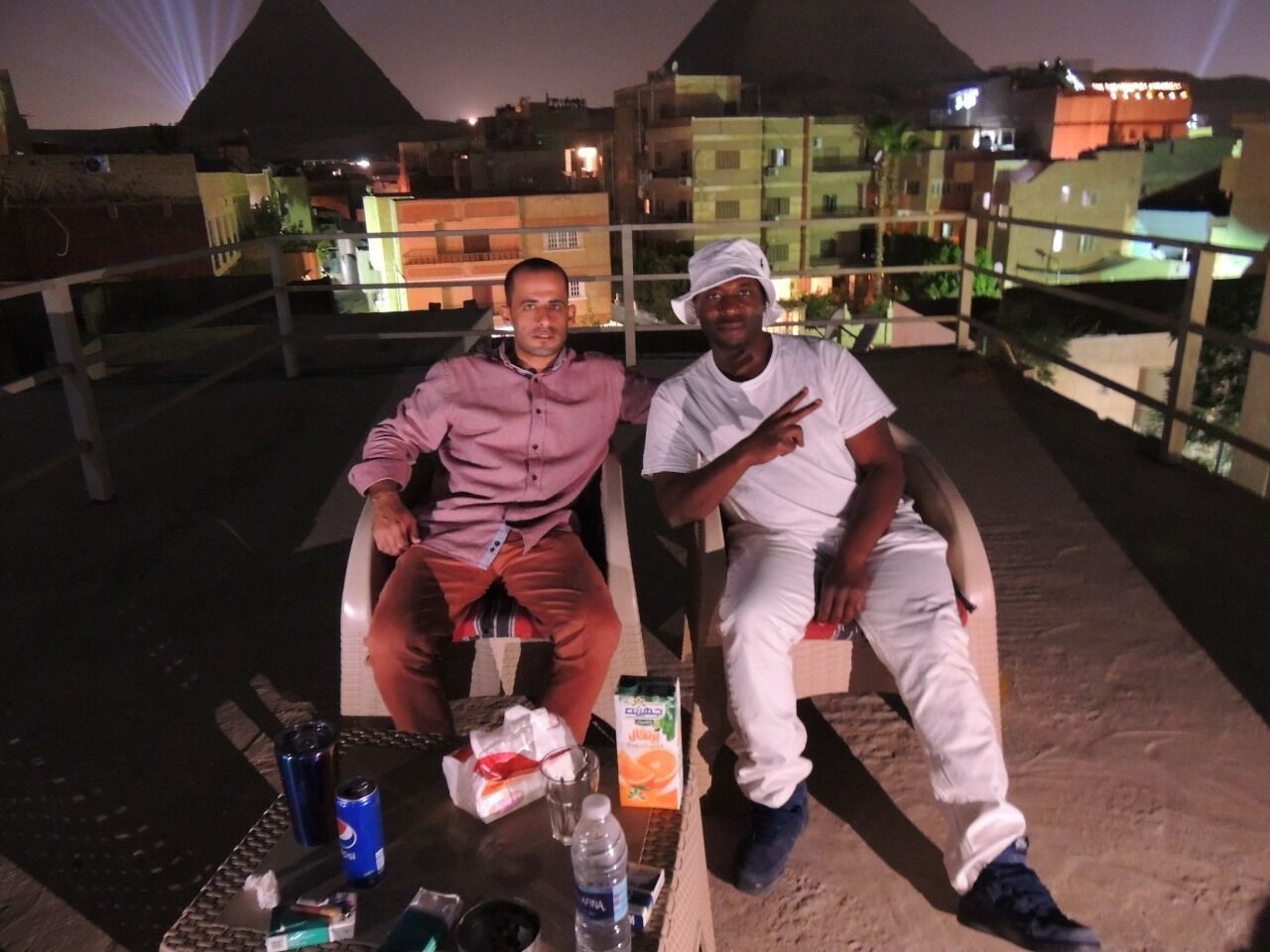 Two men seated at an outdoor table with drinks and snacks, overlooking a cityscape at night.