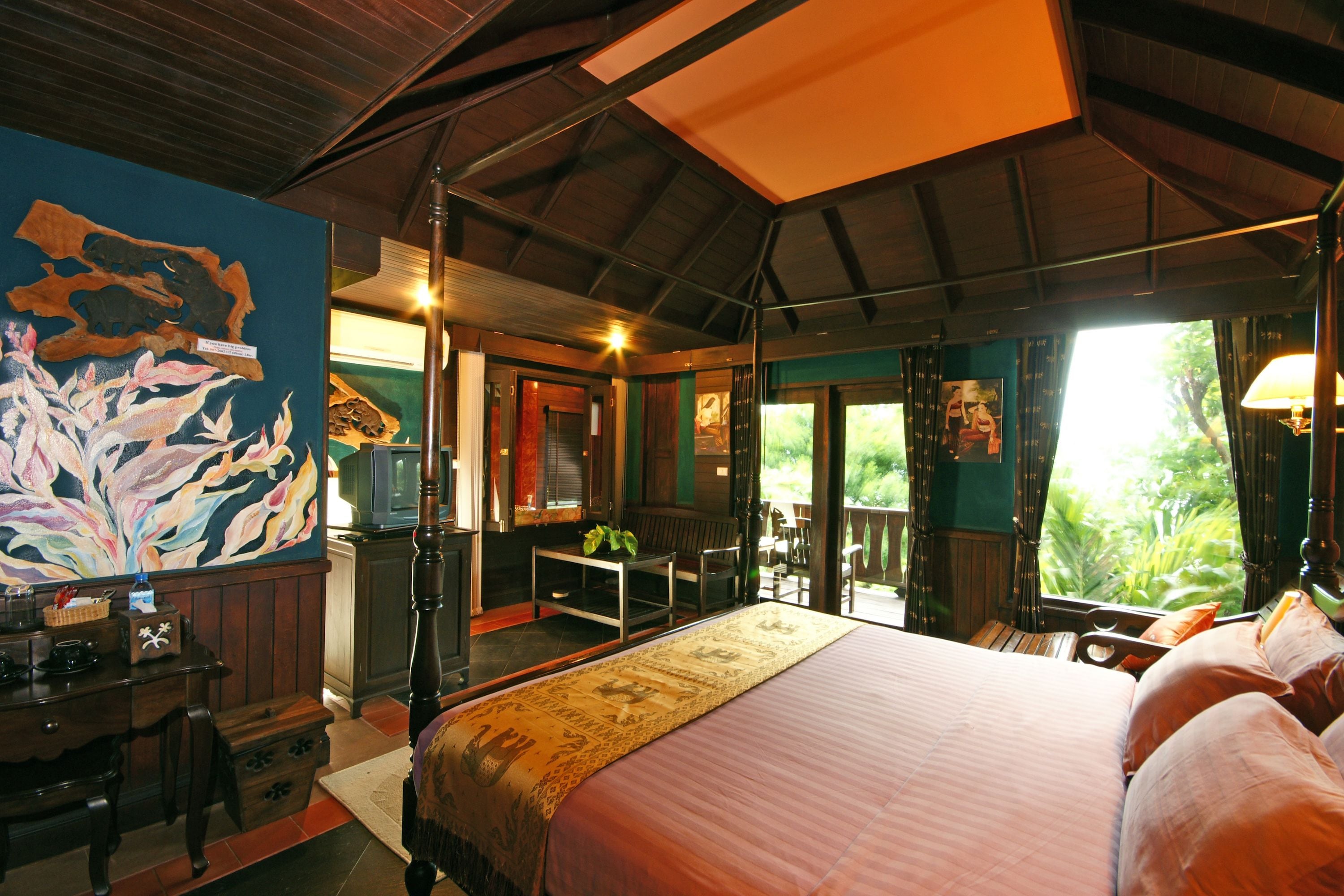 A bedroom with a large bed, a desk, and a television. There is a balcony with a view of greenery.
