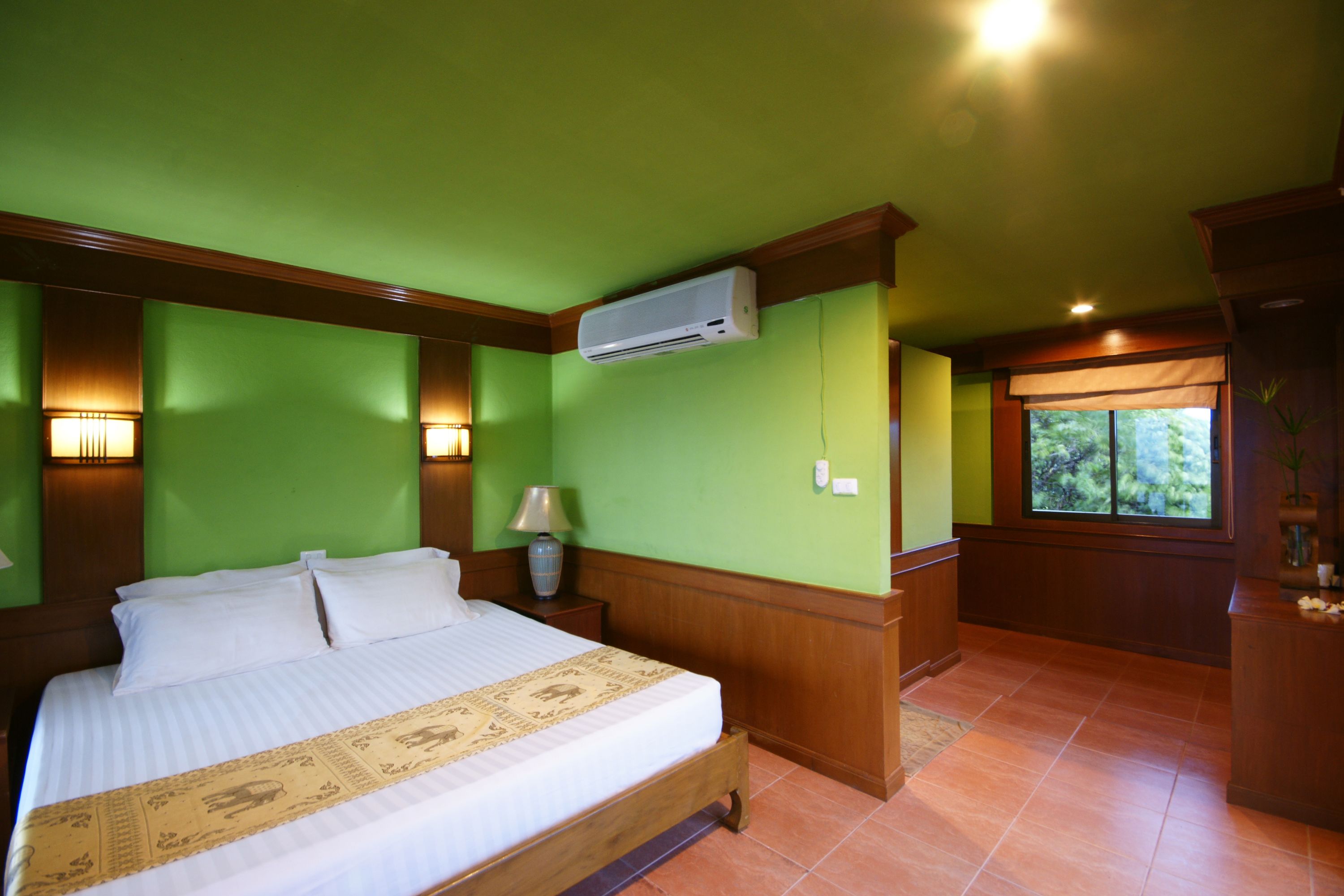 Habitación de hotel con una cama grande, paredes verdes, paneles de madera, un equipo de aire acondicionado, una mesita de noche con lámpara y una ventana con vista a zonas verdes.
