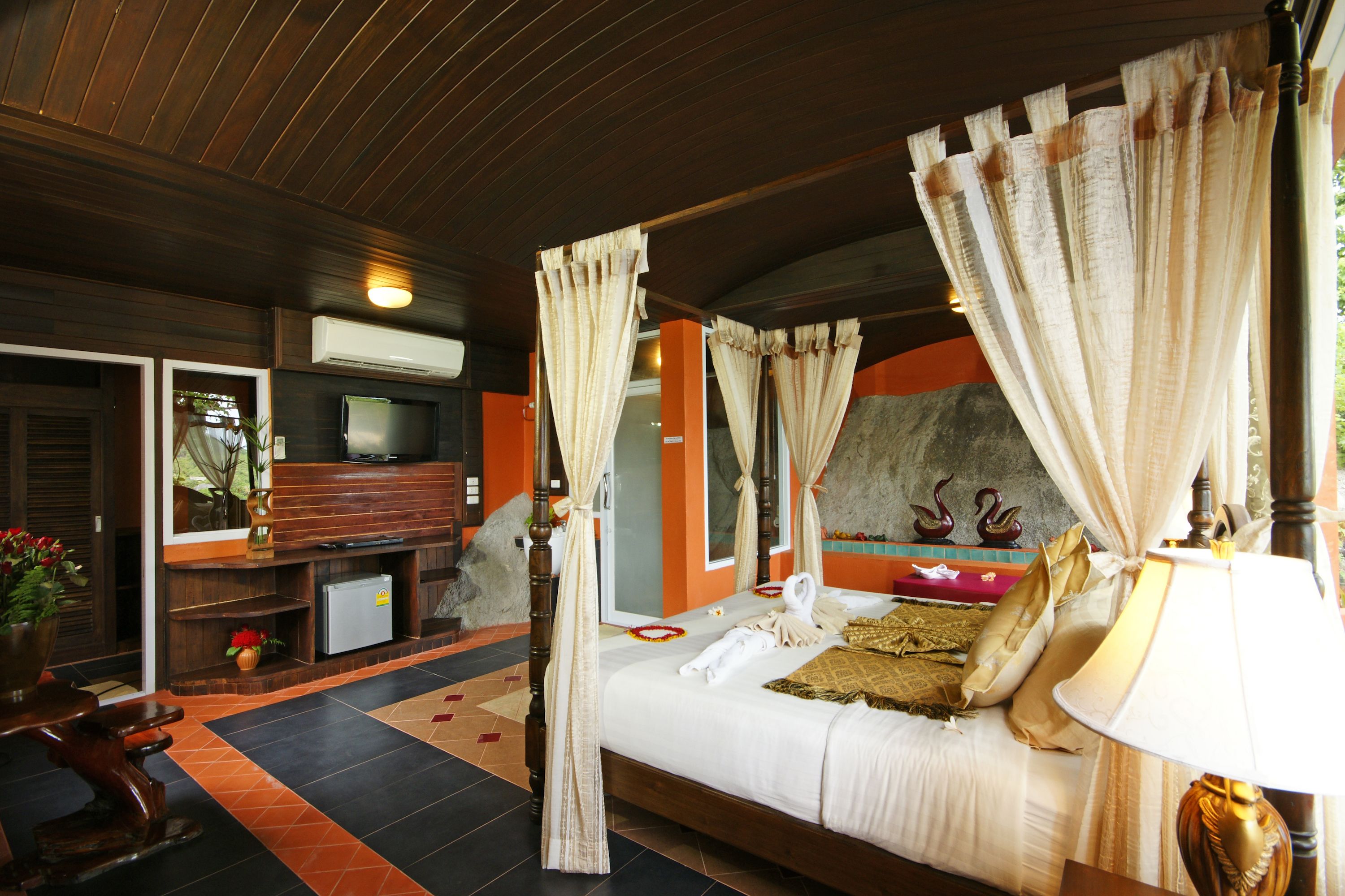 A four-poster bed with curtains, a television, a refrigerator, and a small plant.