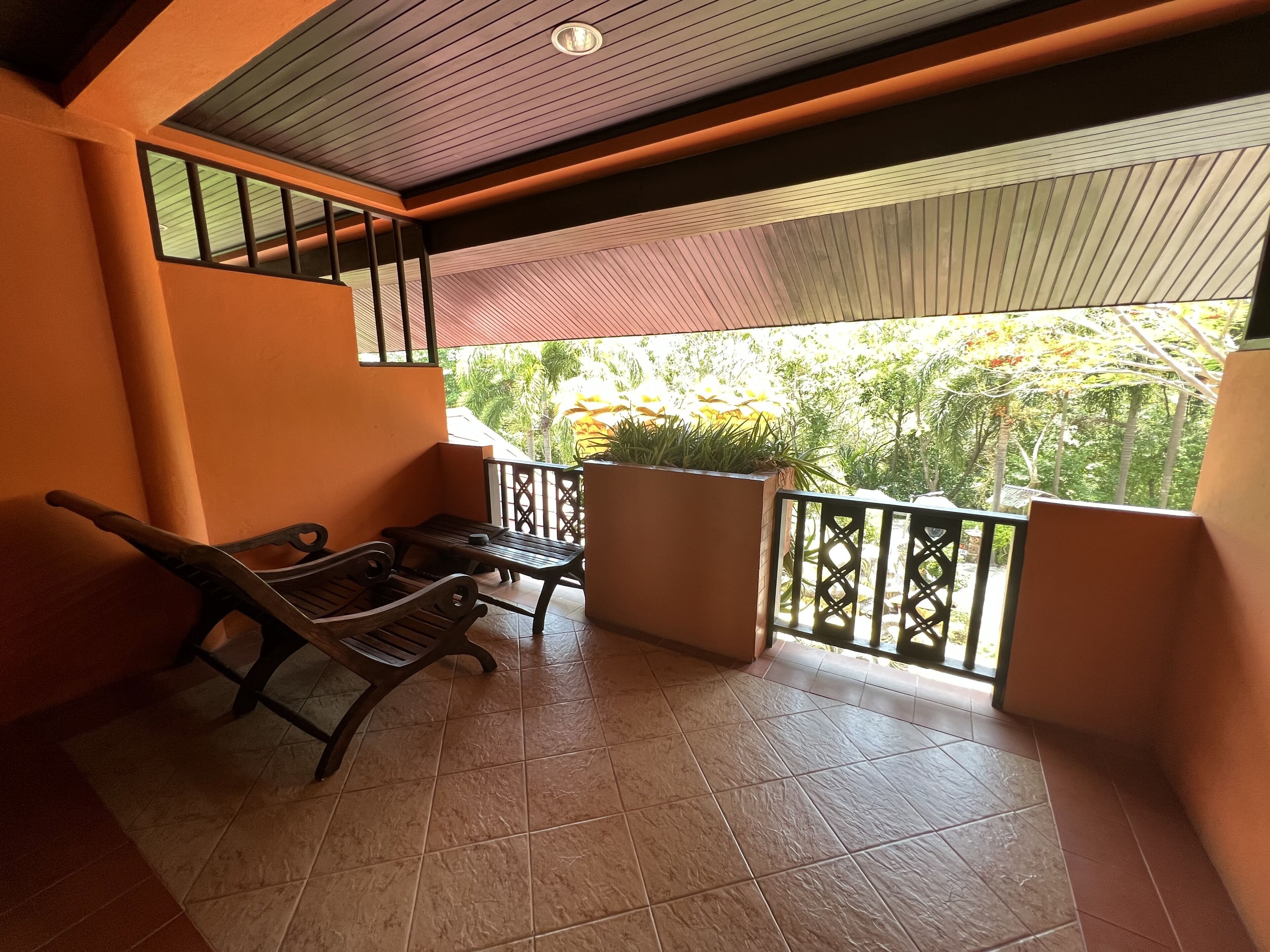 A balcony with a wooden bench and a table, a tiled floor, and a view of greenery outside.