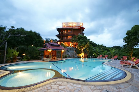A swimming pool with a stone border, a multi-level building with a red roof, and red lounge chairs around the pool.