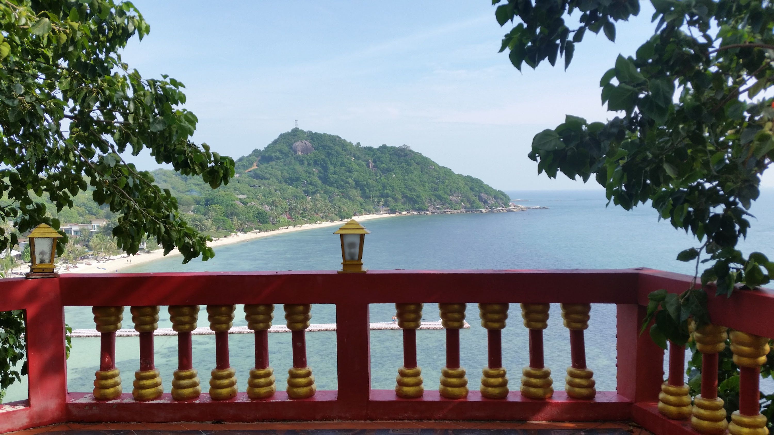 A coastal view from a red balcony with a railing, overlooking a beach and a green hill.