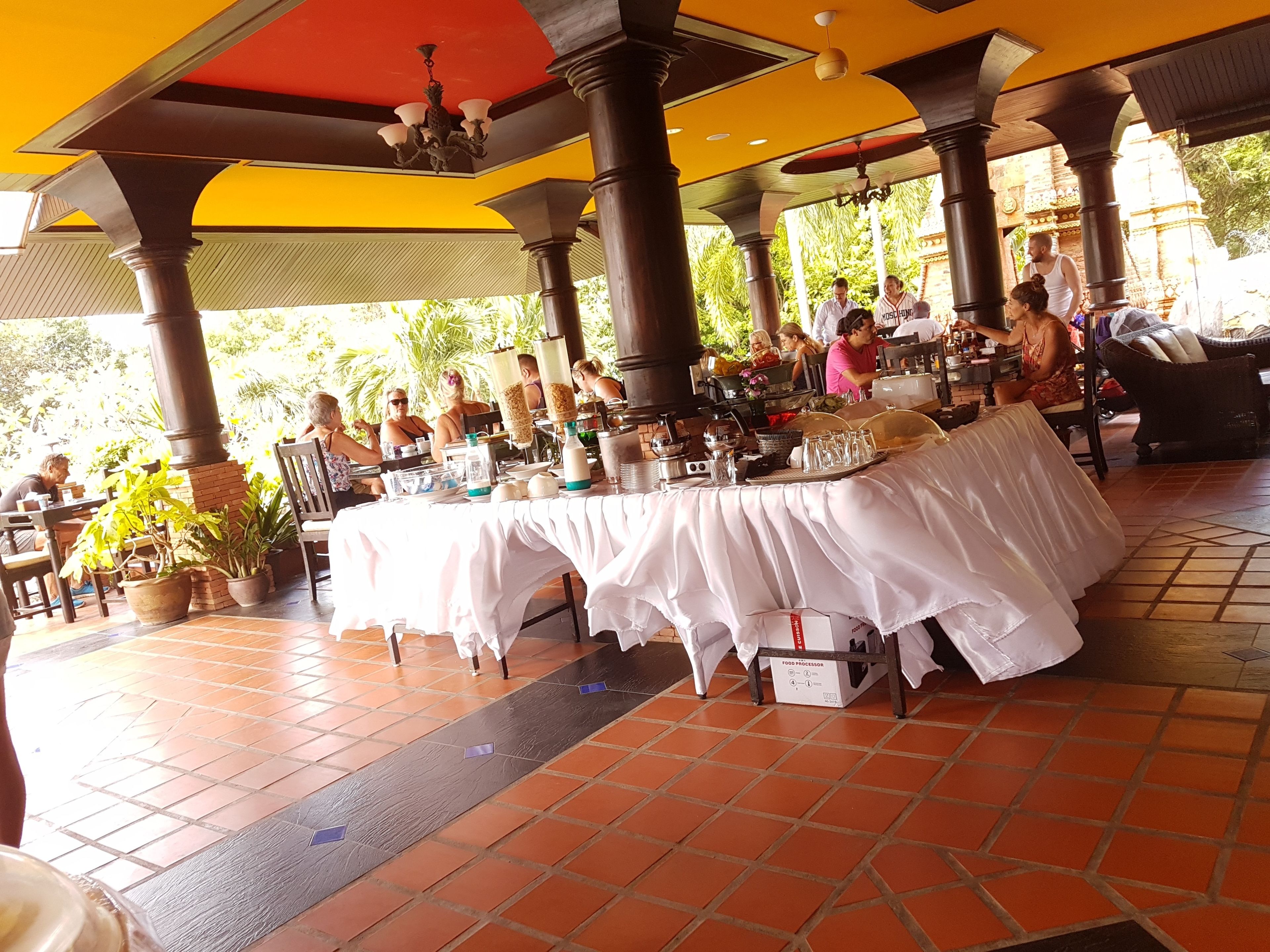 An outdoor dining area with tables covered in white tablecloths, potted plants, and a tiled floor.