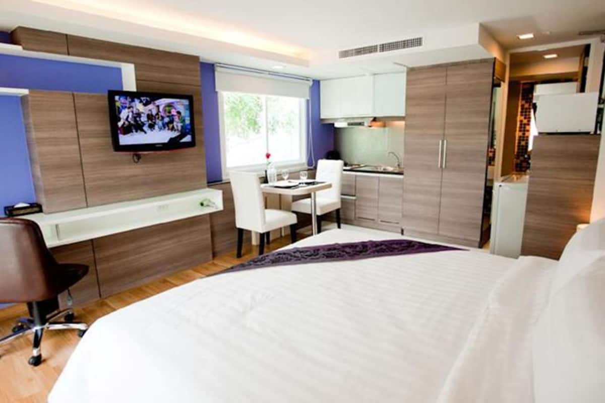 A modern hotel room with a large bed, a desk, a chair, a television, and a kitchenette.