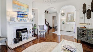 Uma sala de estar espaçosa com lareira, um candelabro e vista para a cozinha através de um arco.