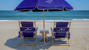 Duas cadeiras de praia e uma mesa sob um guarda-sol azul em uma praia de areia, com o oceano ao fundo.