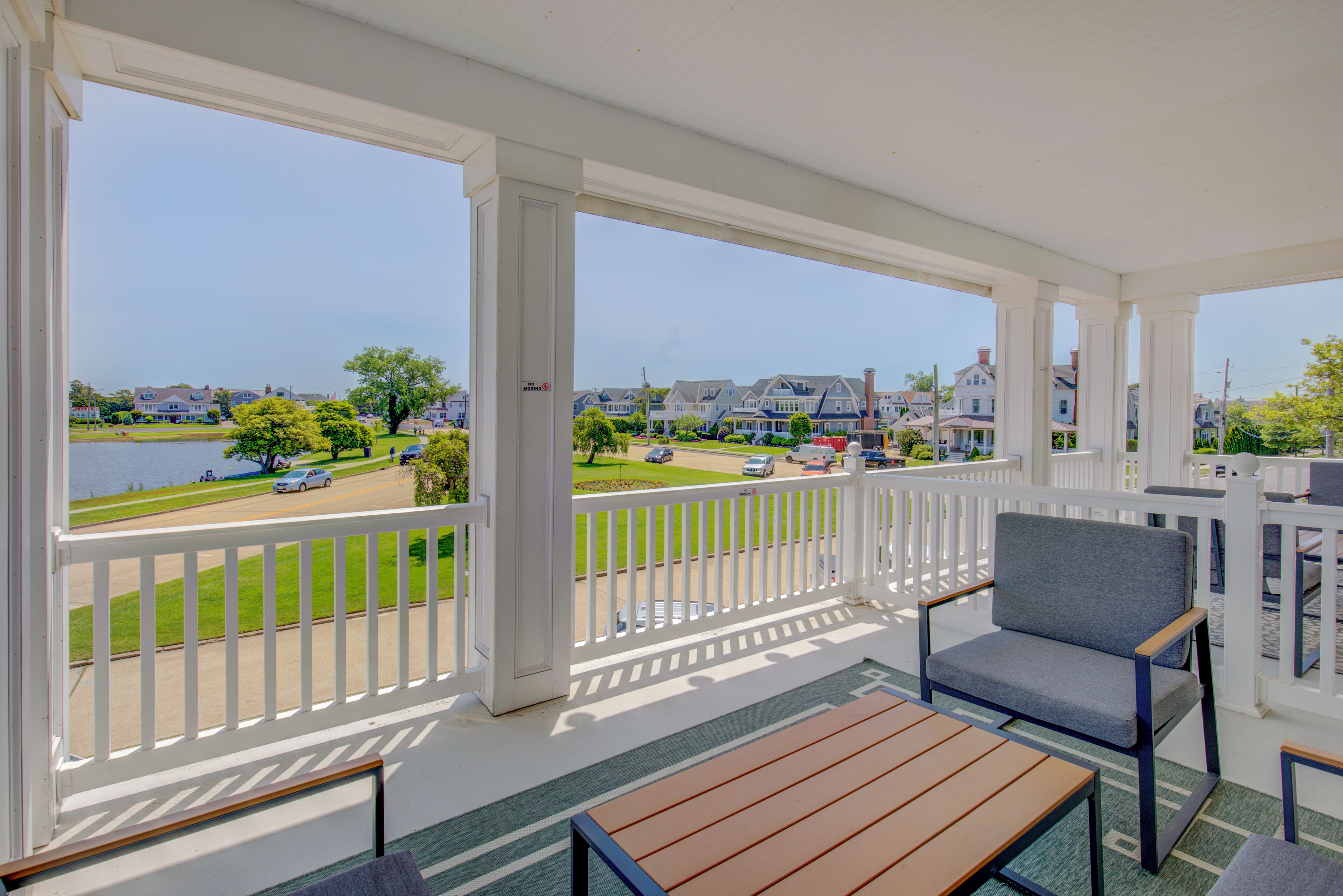 Een overdekt buitengebied met een houten tafel en stoelen, een witte balustrade en uitzicht op een woonstraat met huizen en een waterpartij.