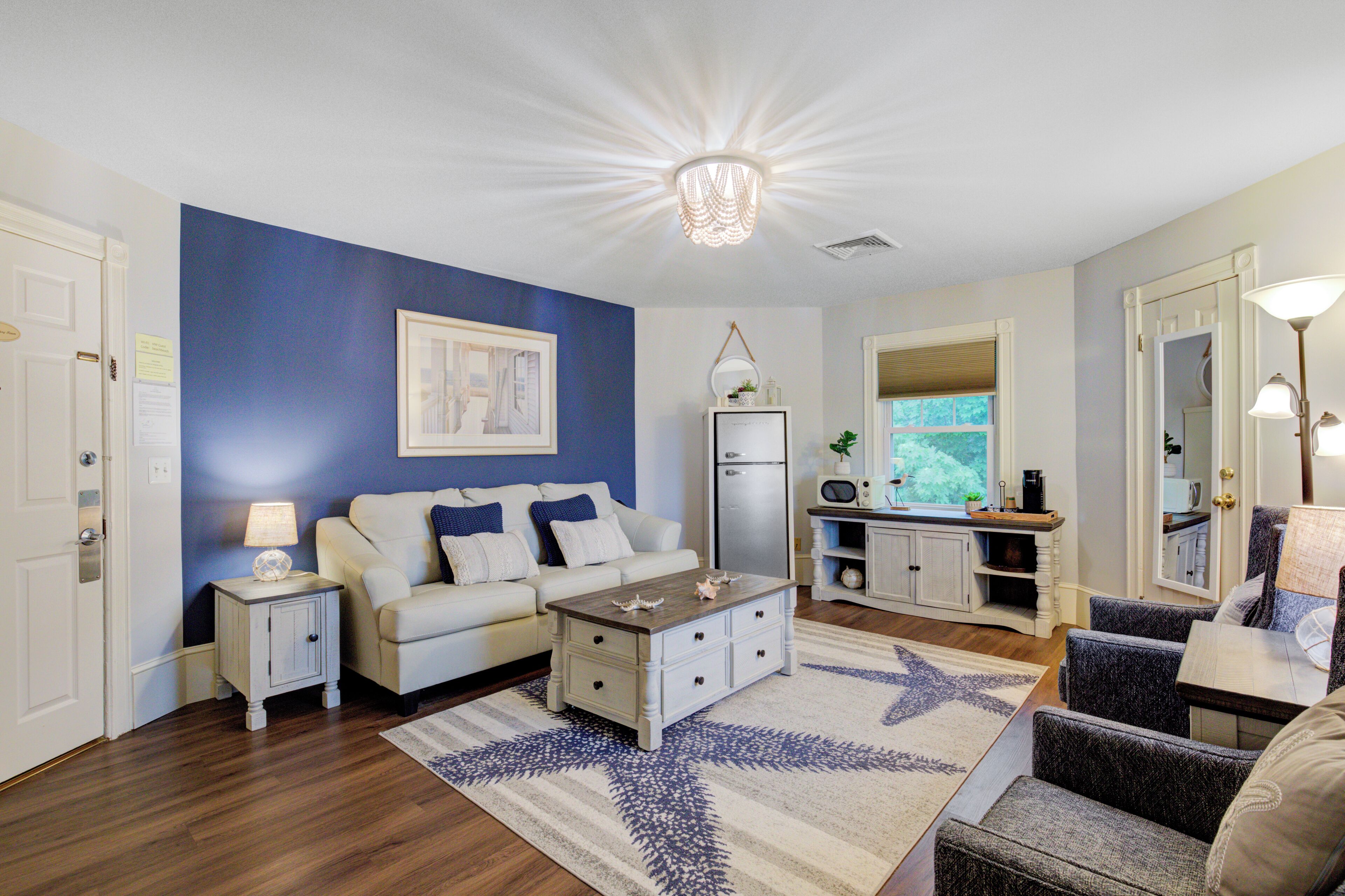 Un salon avec un mur d’accent bleu, un canapé blanc, une table basse et un coin cuisine équipé d’un réfrigérateur et d’un évier.