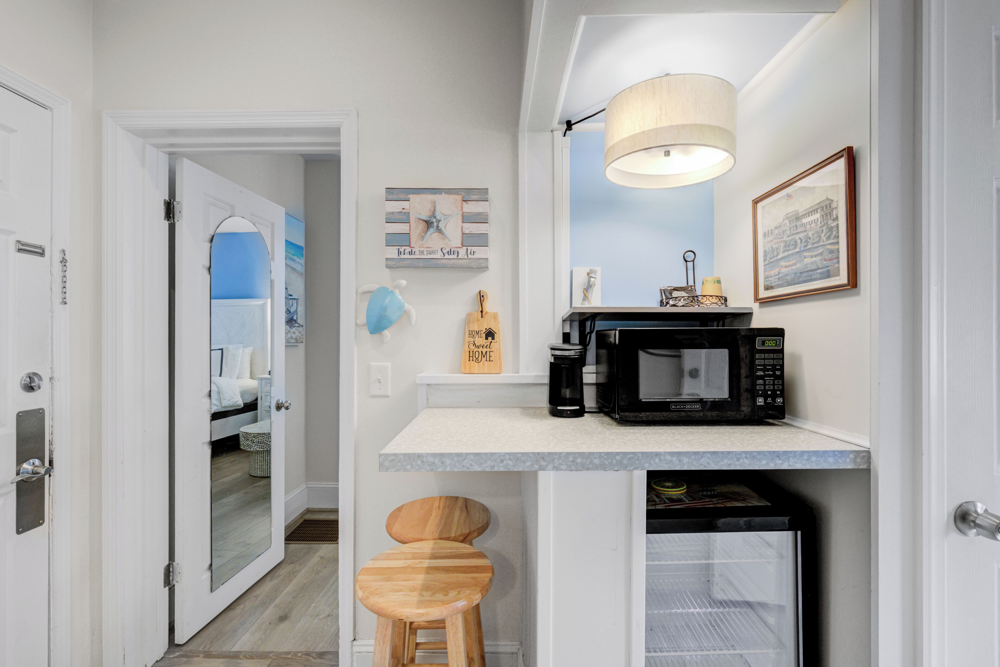 A compact kitchen with a microwave, a coffee maker, and a small refrigerator. There is a wooden stool and a framed picture on the wall.