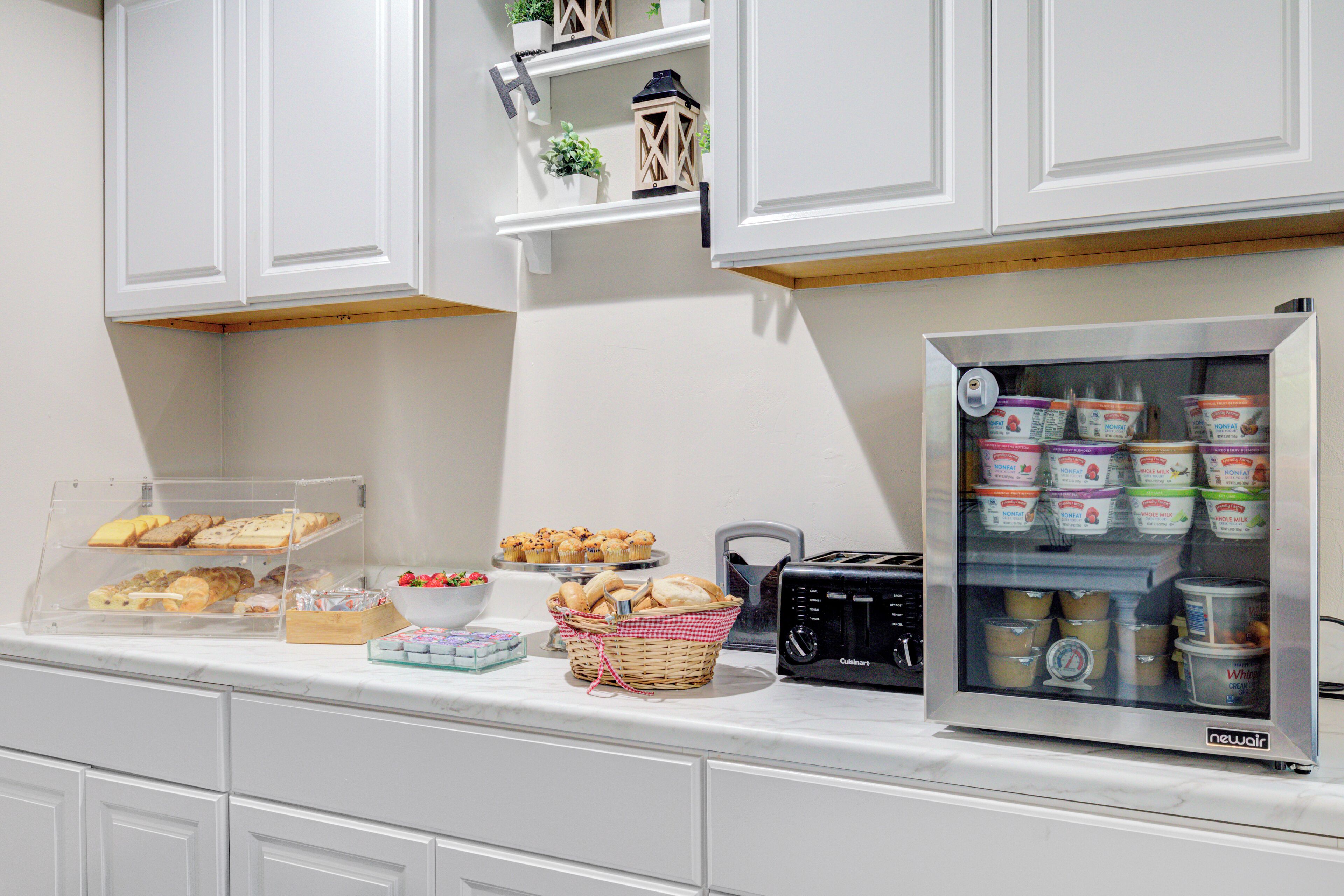 A kitchen counter with a variety of food items, including pastries, bread, and a yogurt refrigerator.