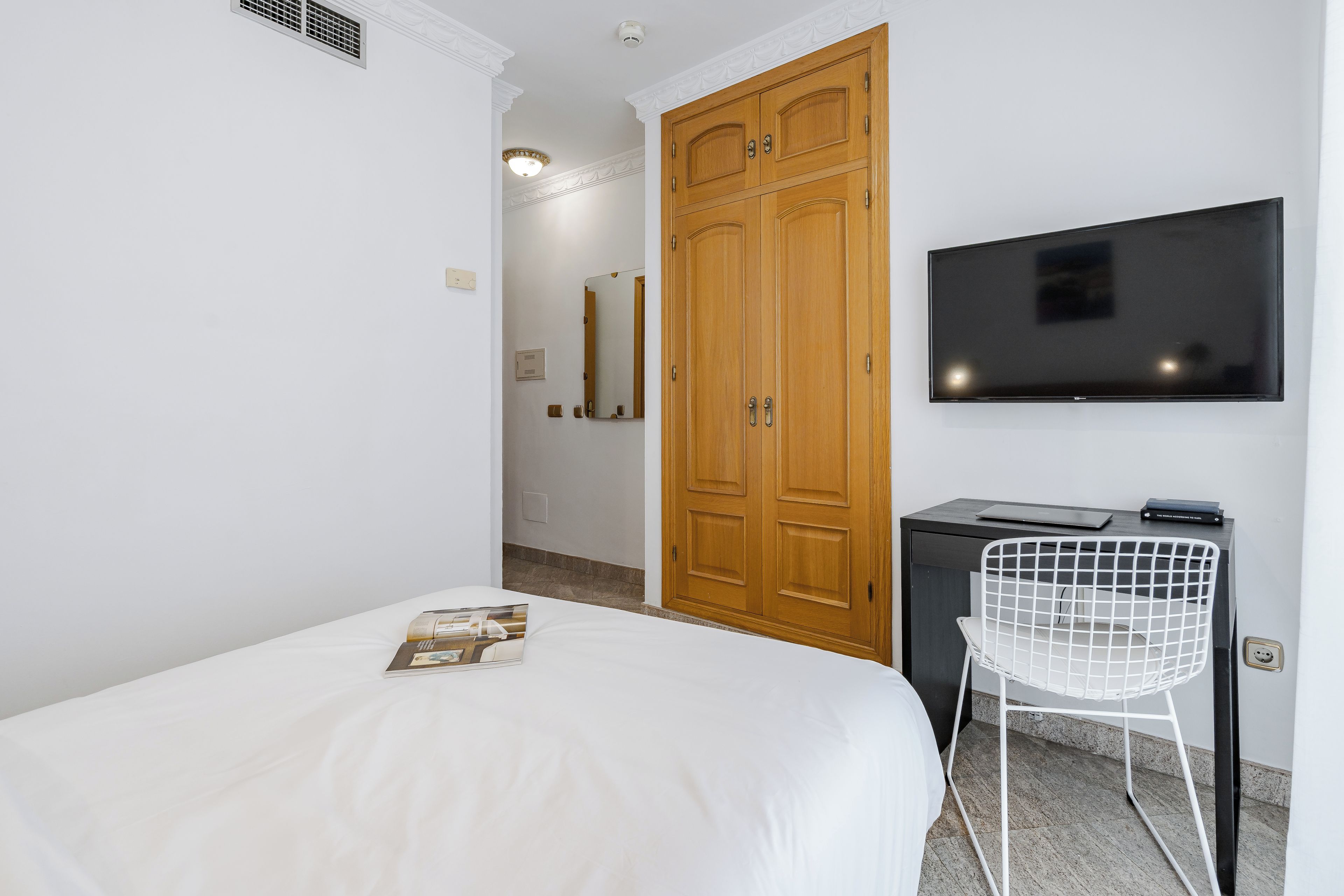 A hotel room with a bed, a TV mounted on the wall, a desk with a chair, and a wardrobe.