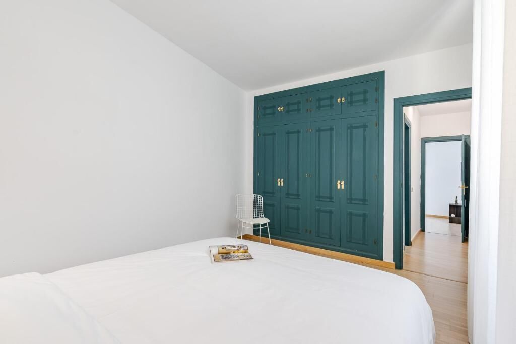 A bedroom with a large bed, a white chair, and a teal wardrobe.