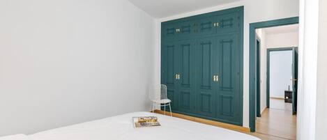 A bedroom with a large bed, a white chair, and a teal wardrobe.
