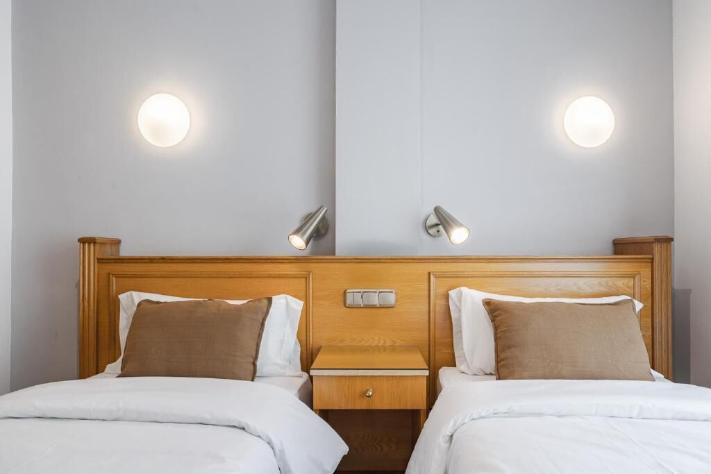 A hotel room with two beds, each with white bedding and brown pillows. There are two bedside tables with lamps, and a wooden headboard with a mirror above.