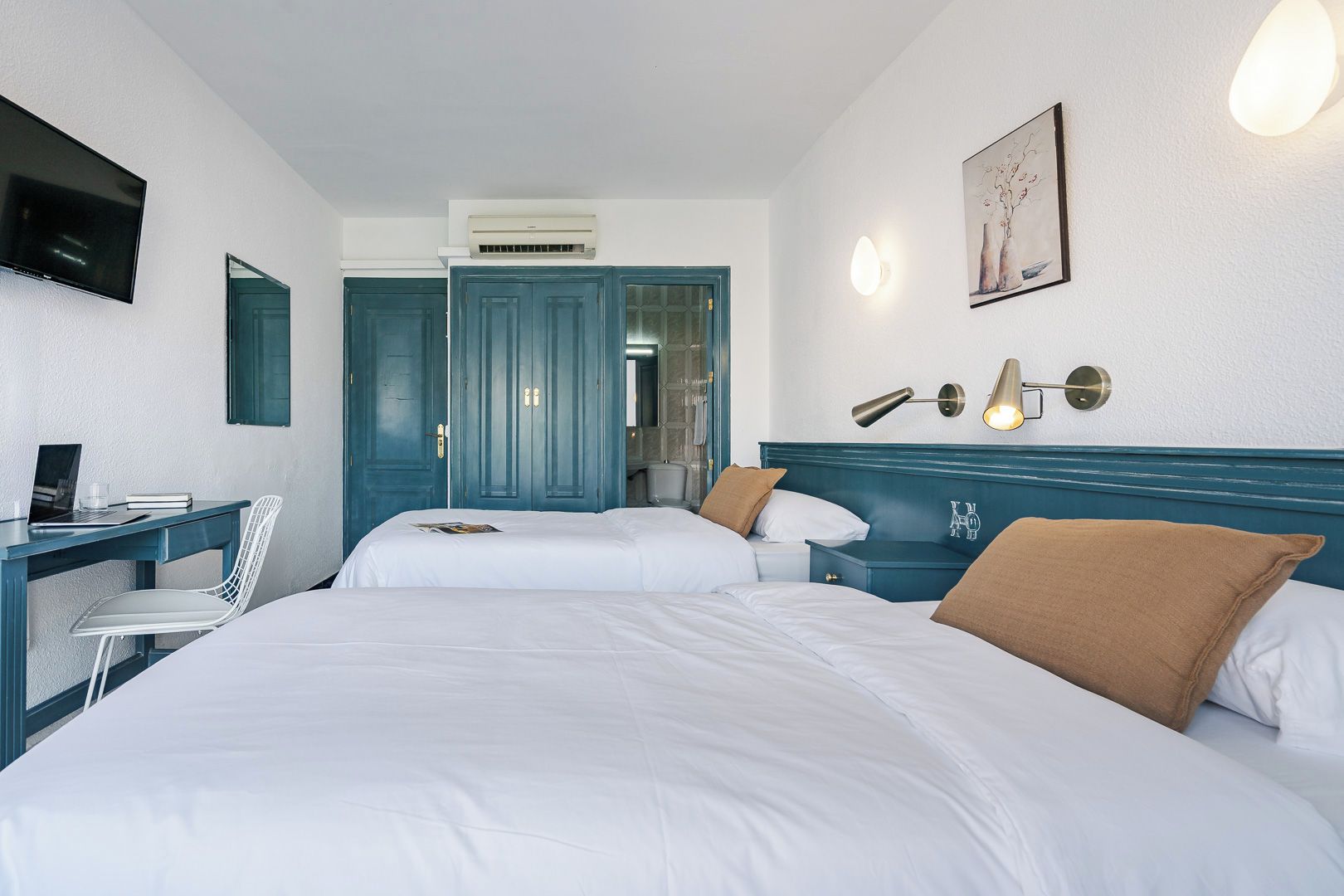 A hotel room with two beds, a desk with a laptop, a chair, a TV, and a wall-mounted light fixture.