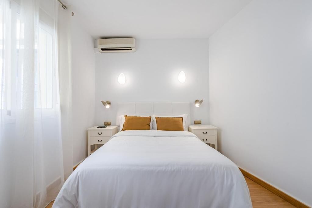 A hotel room with a bed, white walls, a ceiling air conditioner, two bedside tables with lamps, and a window with sheer curtains.