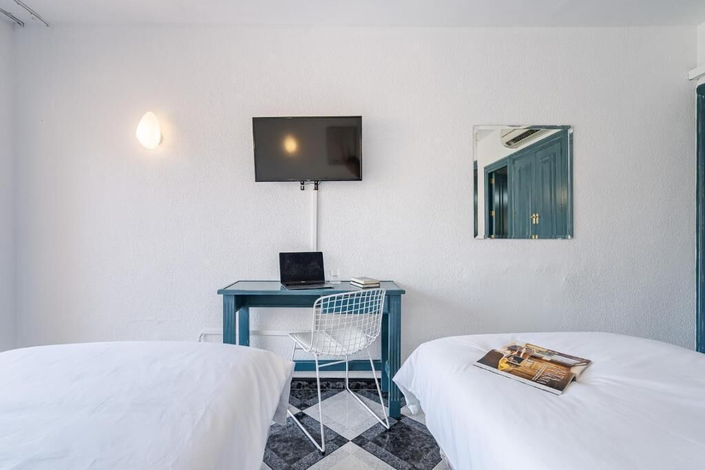 A hotel room with two beds, a desk with a laptop, a chair, a TV mounted on the wall, and a mirror reflecting a door.