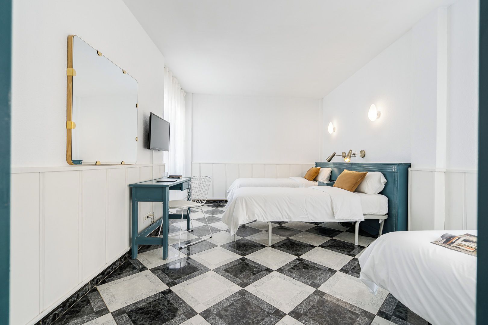 Ein Hotelzimmer mit Schachbrettmuster-Boden, einem Schreibtisch mit Stuhl, zwei Betten mit Kopfteilen, einem Fernseher und Wandleuchten.