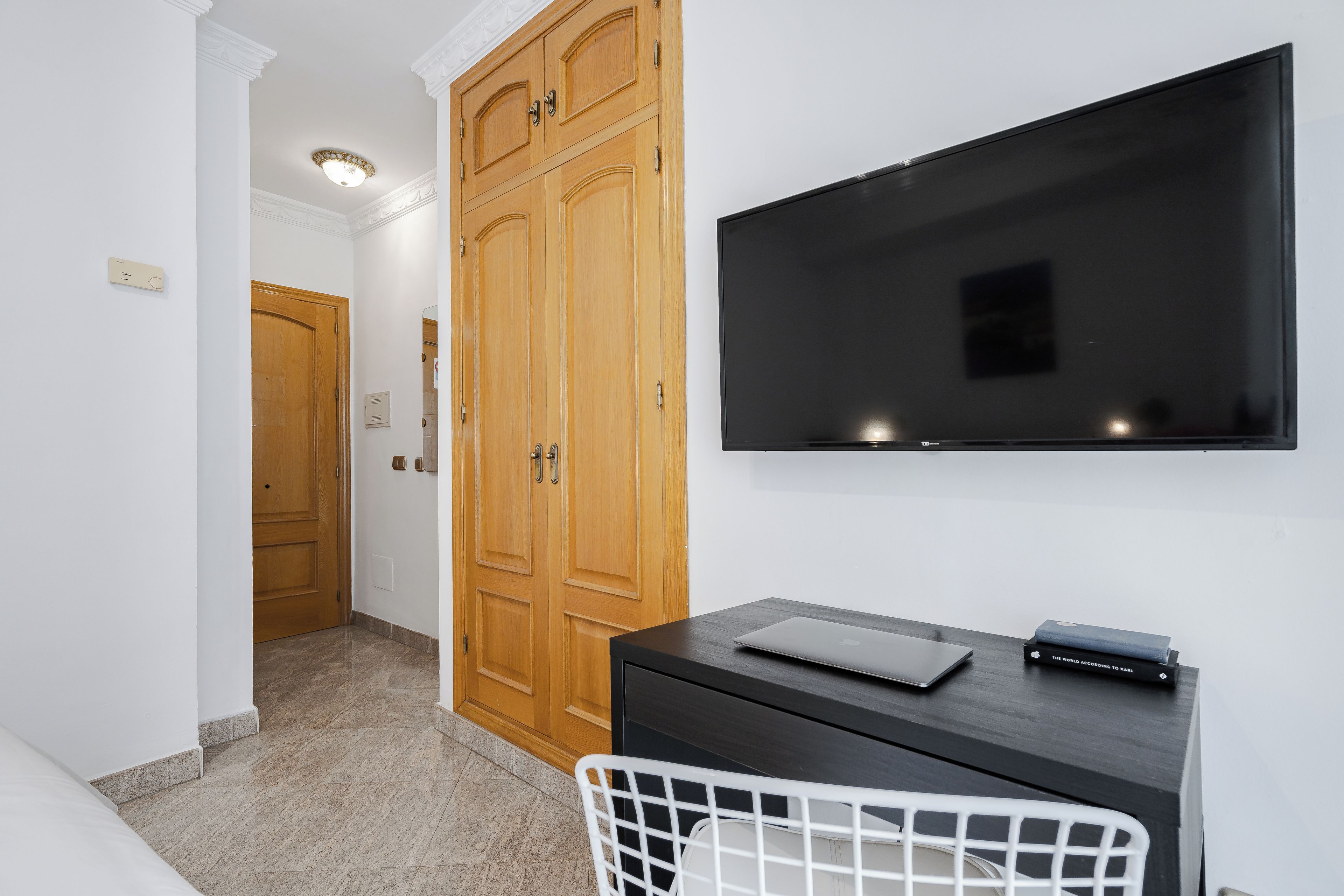 Habitación de hotel con armario de madera, televisor de pantalla plana en la pared, escritorio con una computadora portátil y una silla blanca.
