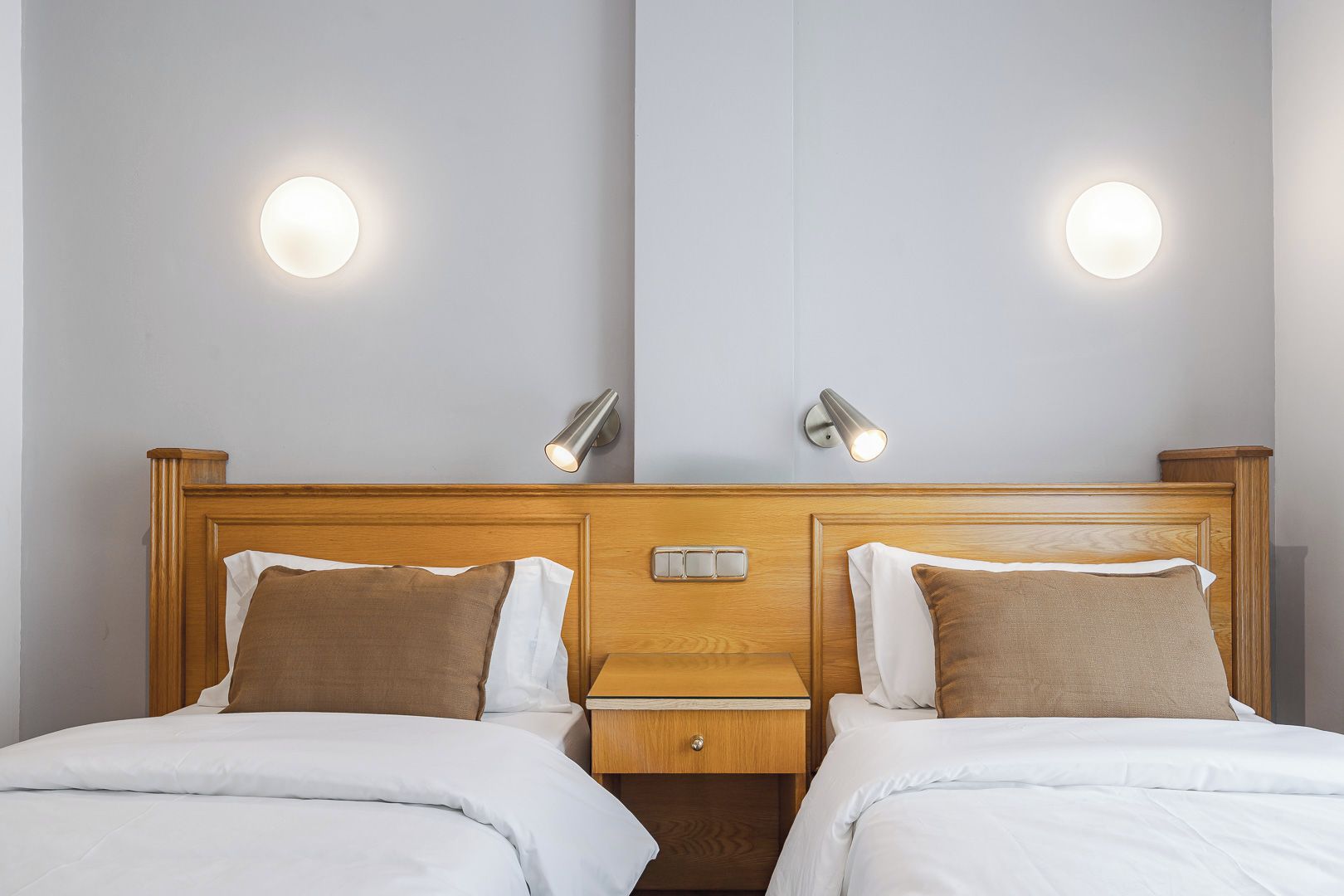 A hotel room with a wooden headboard, two bedside tables, a nightstand, and two round wall sconces.