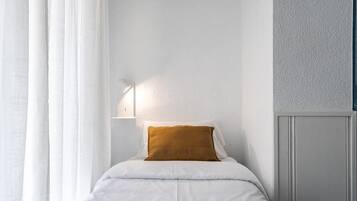 Una cama bien hecha con ropa de cama blanca y una almohada color mostaza, una mesita de noche fijada a la pared con una lámpara, y una pared lisa con una sección texturizada.
