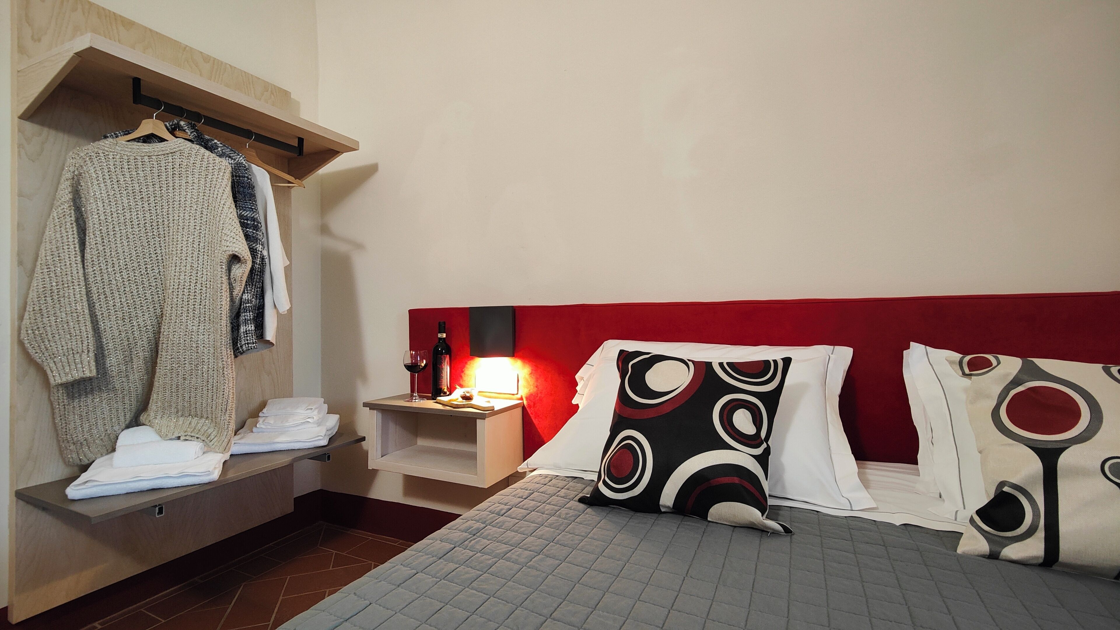A hotel room with a bed, a bedside table with a lamp, a hanging robe, and a shelf with folded towels.
