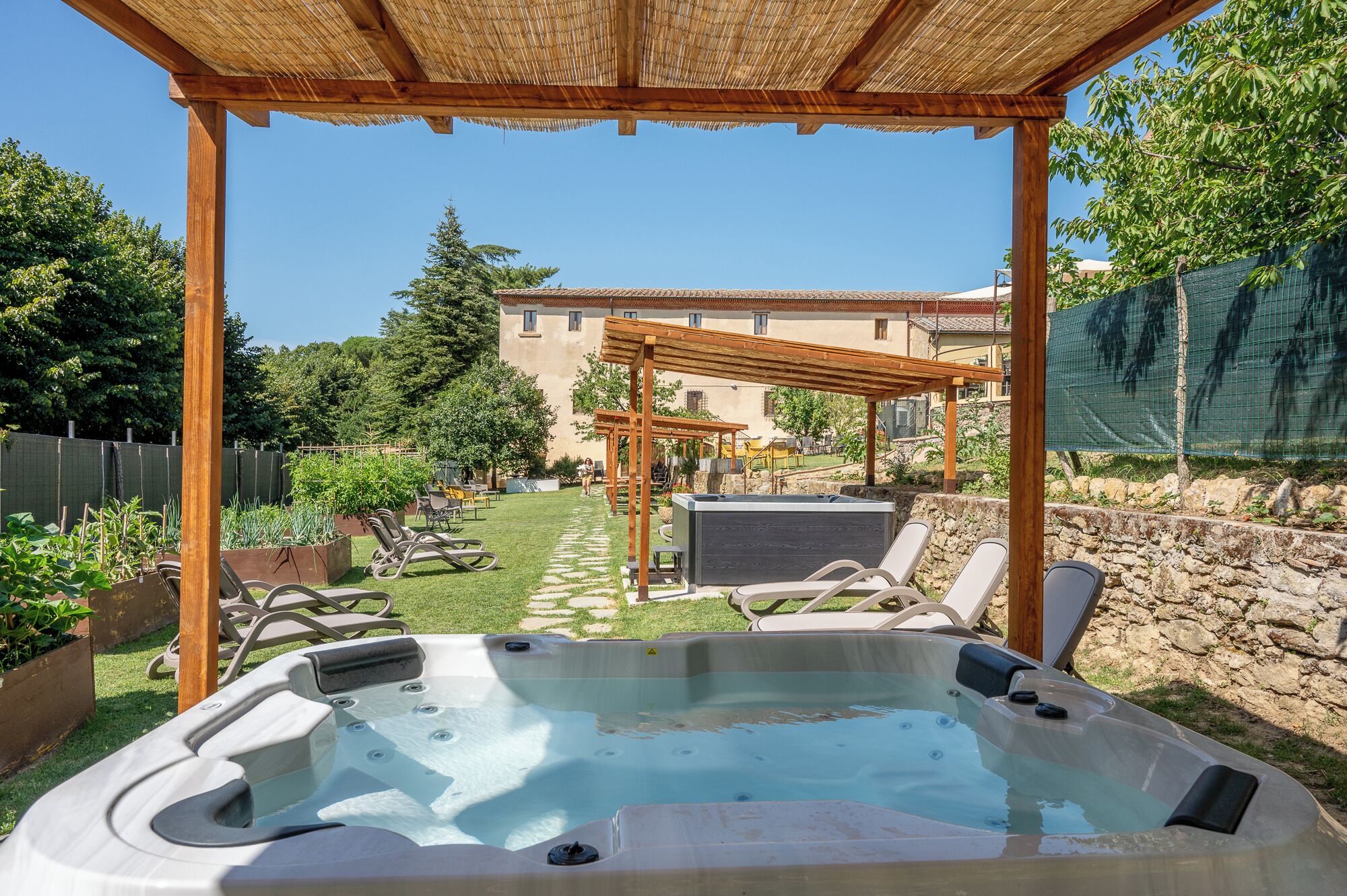 A hot tub under a wooden pergola, a garden with chairs, and a building in the background.