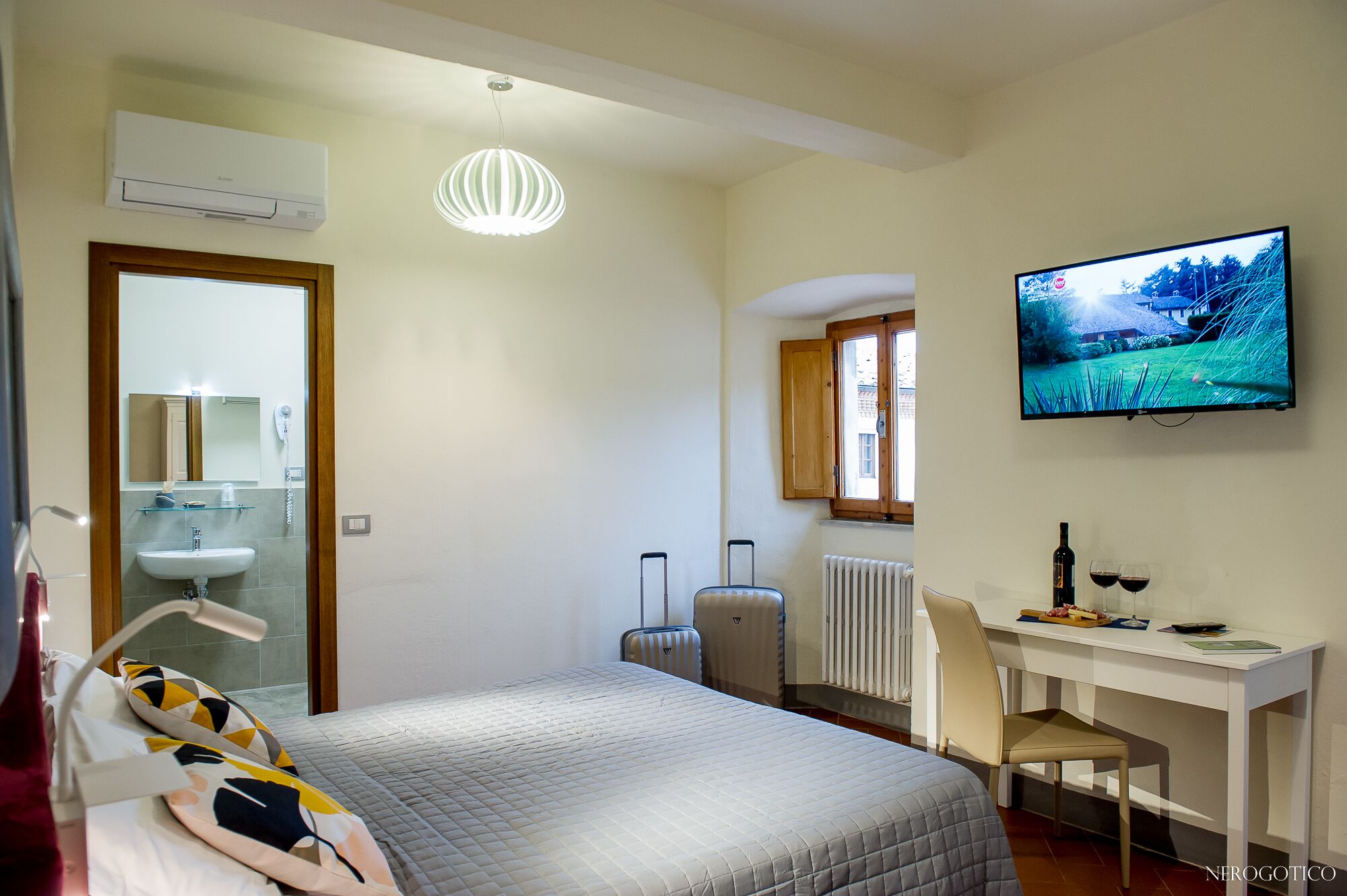 Una habitación de hotel con cama, escritorio con silla, televisor, radiador, ventana y baño con lavabo y espejo.