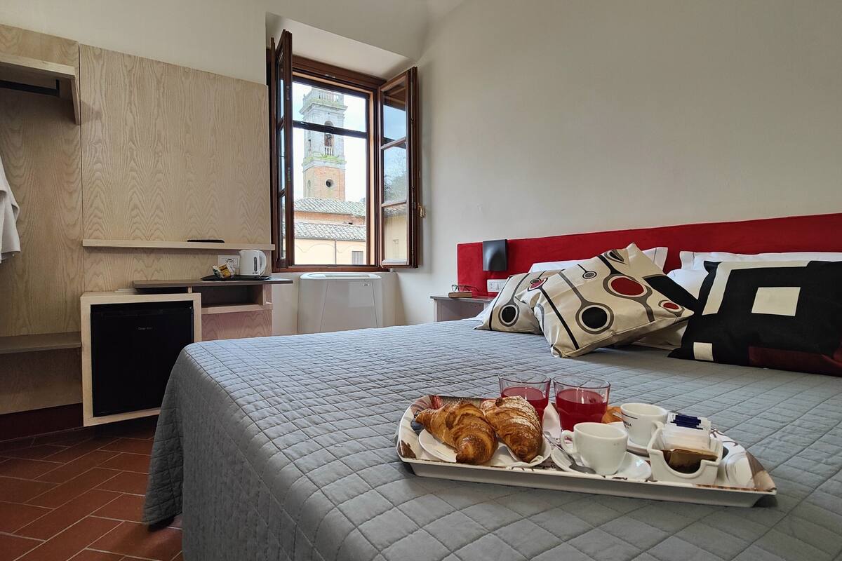 Ett hotellrum med en säng, ett brickbord med frukost och utsikt över en byggnad genom ett fönster.